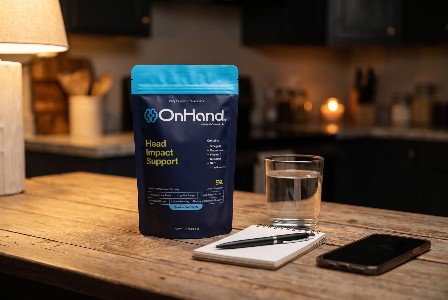 The OnHand pouch standing on a kitchen table at night beside a glass of water, a blank notepad with a pen and a phone face down, under warm lamp light