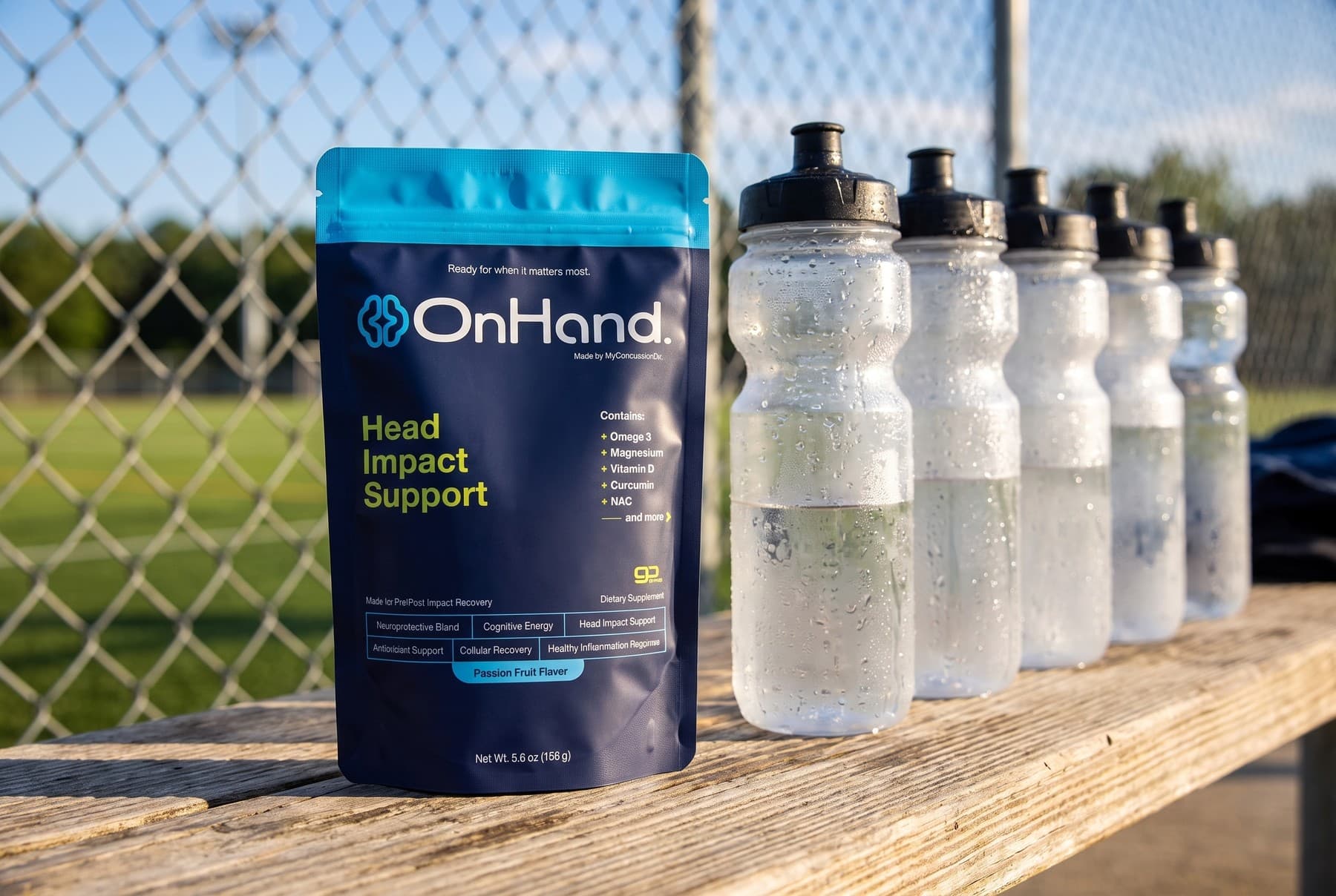 The OnHand pouch standing on a weathered wooden bench beside a row of clear sports water bottles beaded with condensation, a grass field and fence behind in bright afternoon light